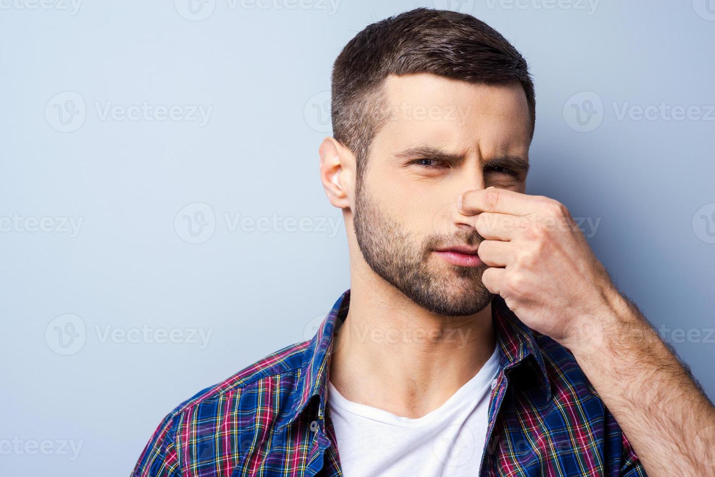 Disgusting smell. Portrait of frustrated young man in casual shirt holding nose and expressing negativity while standing against grey background photo