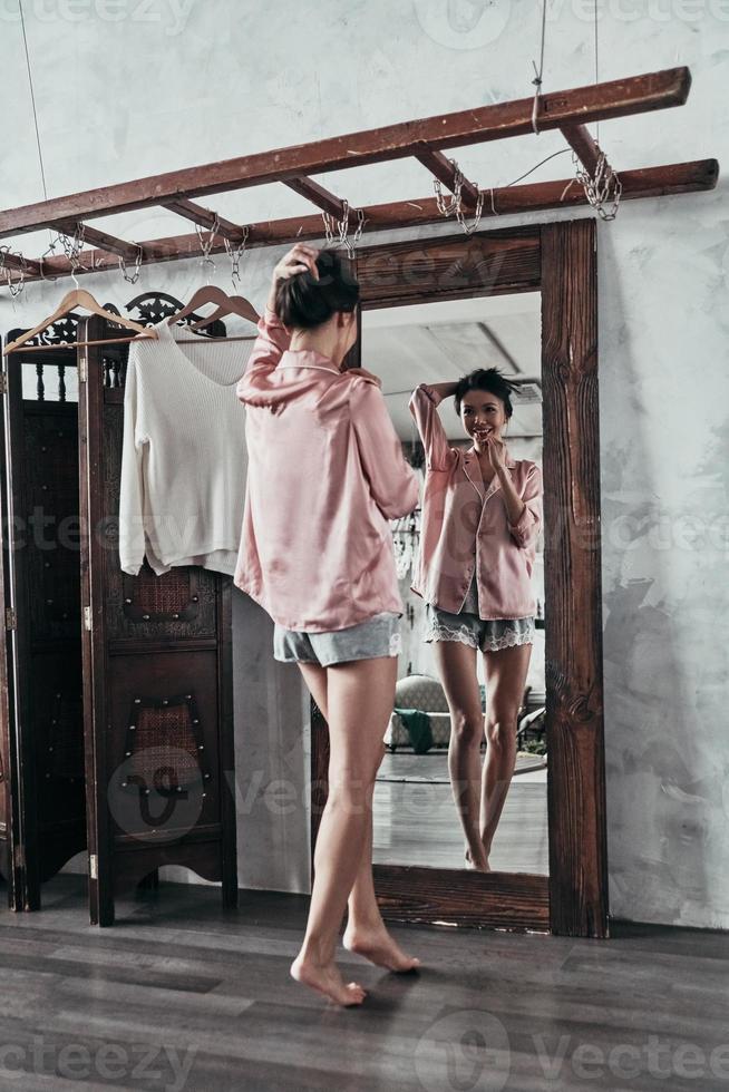 pura belleza femenina. longitud completa de una joven atractiva que se mantiene la mano en el cabello y sonríe mientras está de pie frente al espejo en casa foto