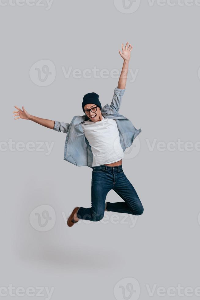 hombre en el aire. toda la longitud de un apuesto joven con camisa de jeans que mantiene los brazos extendidos y mira la cámara con una sonrisa mientras salta contra el fondo gris foto