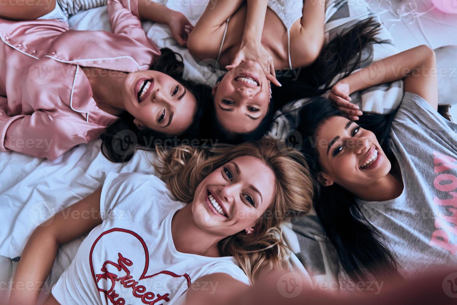 True feminine beauty. Top view self portrait of four attractive young women in pajamas smiling and looking at camera while lying on the bed at home photo