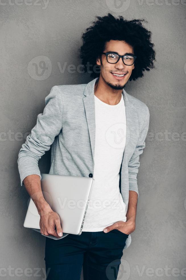 Keep on smiling. Cheerful young African man holding laptop and looking at camera with smile while standing against grey background photo