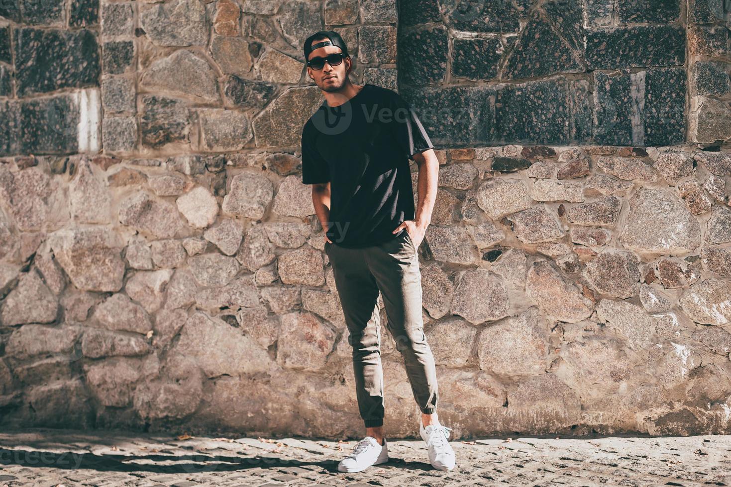 genial y guapo. toda la longitud de un apuesto joven africano con ropa informal parado contra la pared empedrada al aire libre foto