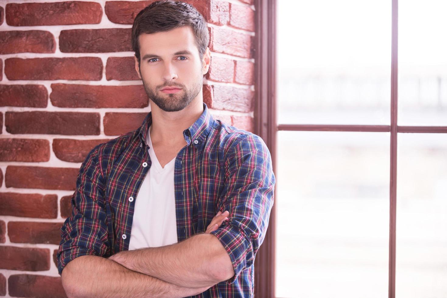 Charming and confident. Handsome young man keeping arms crossed and looking at camera while standing indoors photo