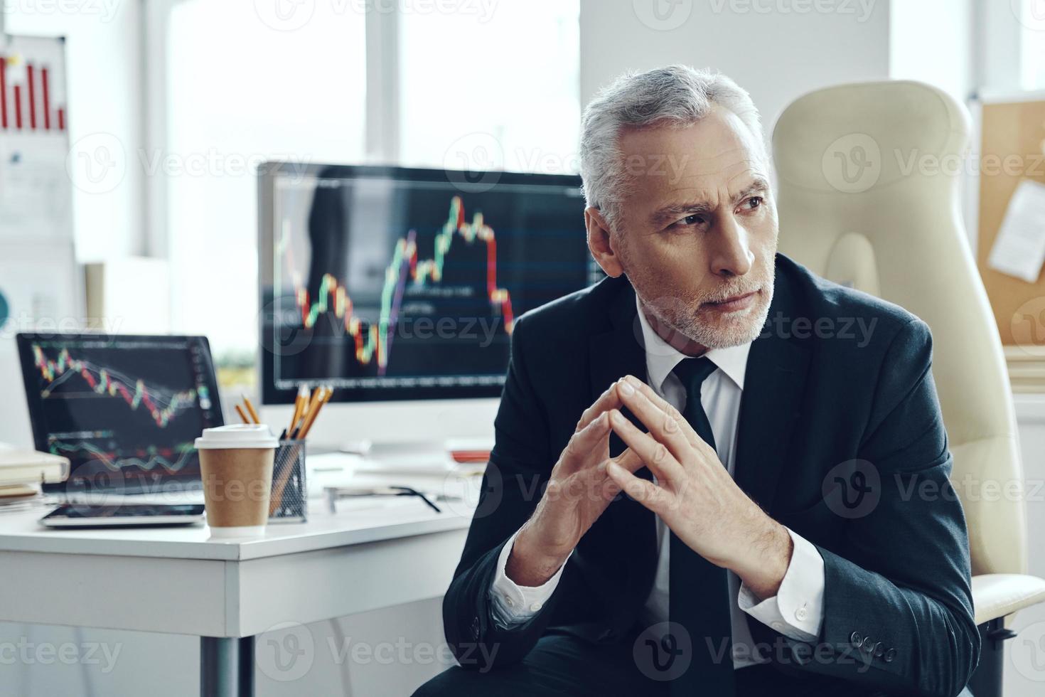 Senior trader in elegant business looking away and keeping hands clasped while working at the office photo