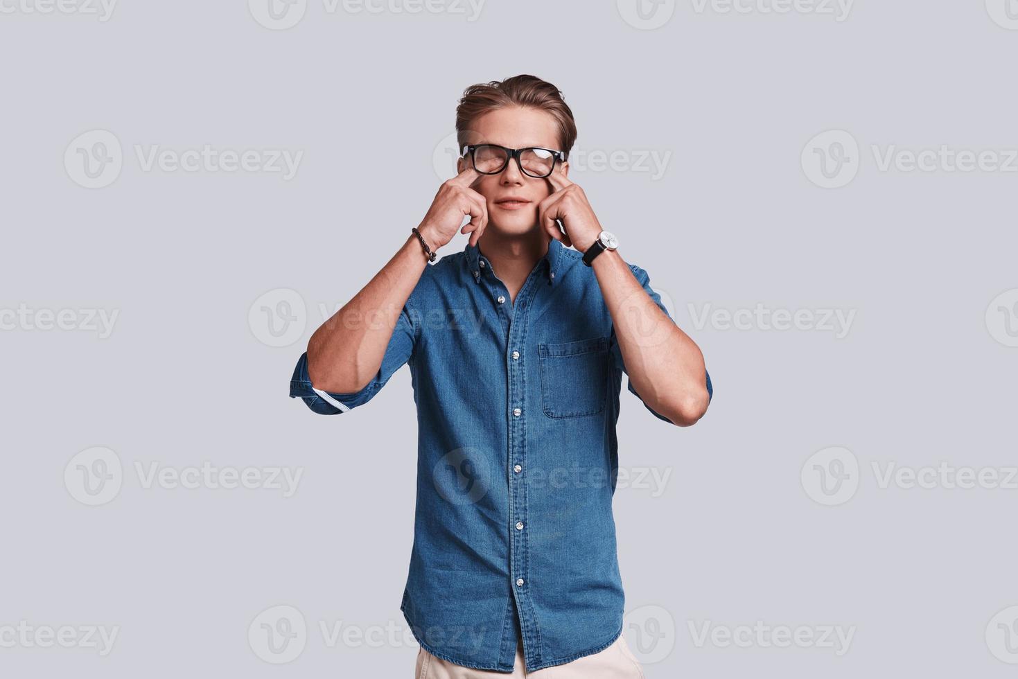 Just a little tired.  Handsome young man touching his eyes with fingers while standing against grey background photo