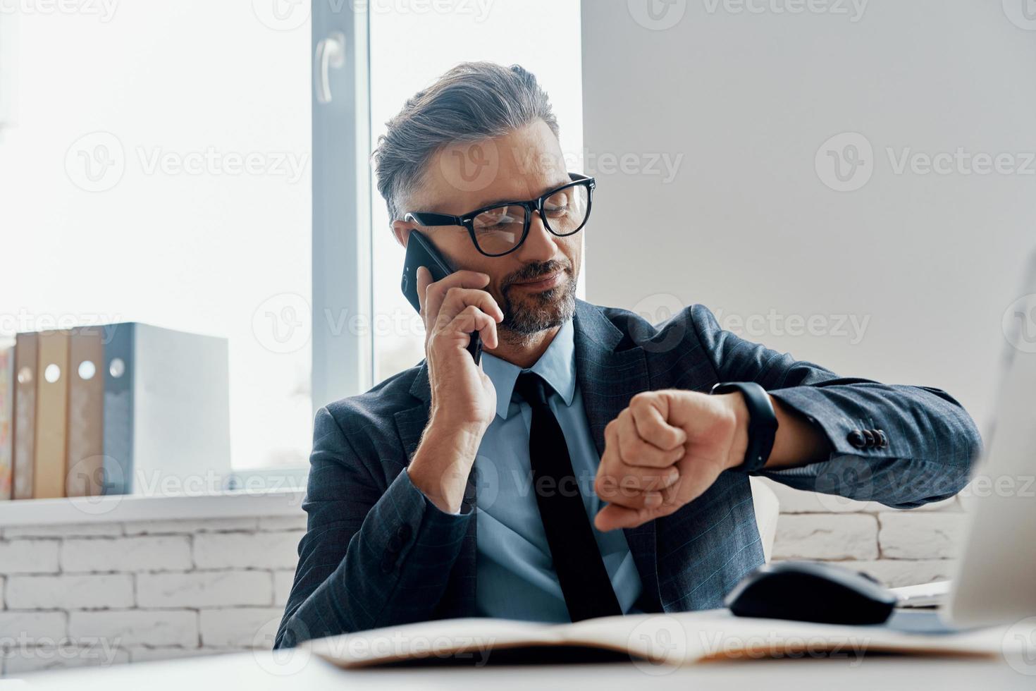 Handsome mature man in formalwear checking the time while talking on mobile phone in the office photo