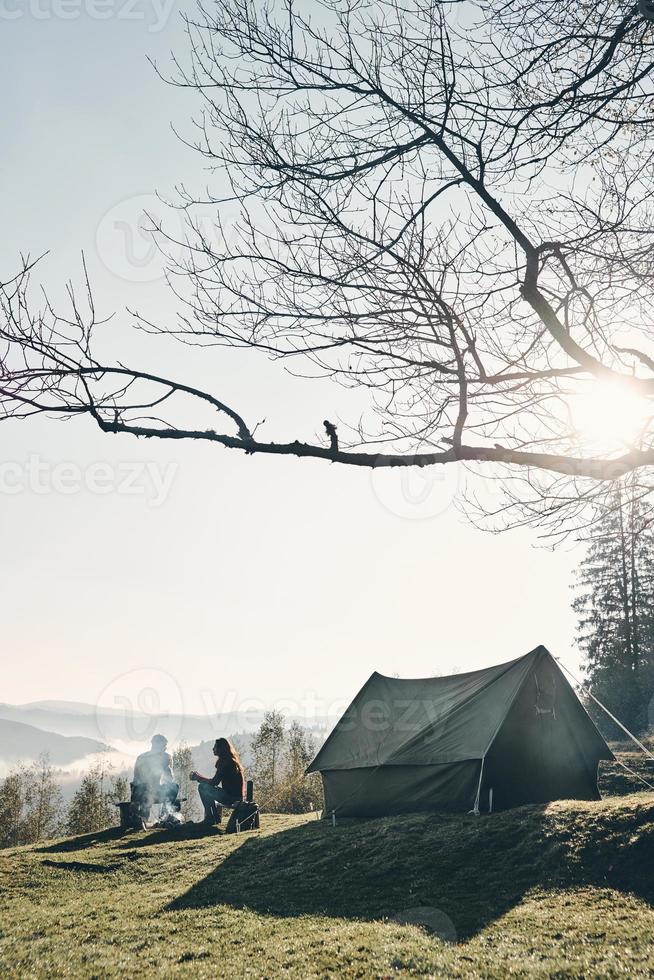 Fresh air and perfect view. Young couple having morning coffee while sitting near the tent in mountains photo