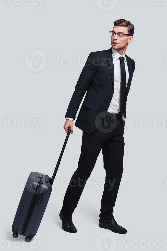In a hurry. Full length of good looking young man with suitcase looking away while standing against grey background photo