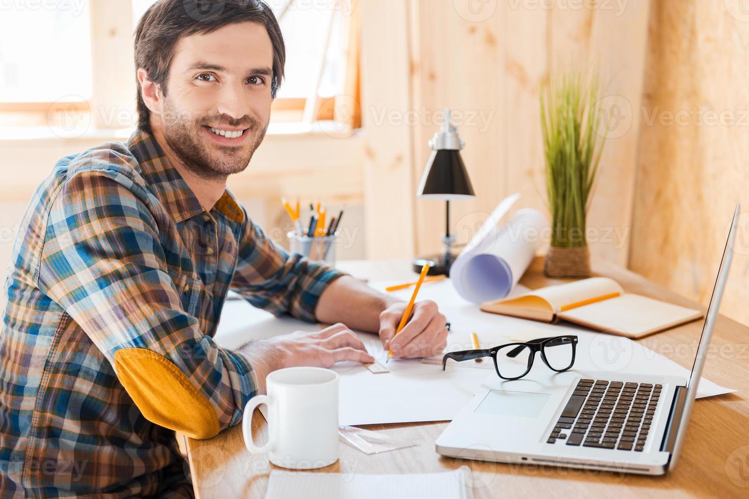 Realizing his ideas. Side view of smiling young man drawing and looking at camera while sitting at his working place photo