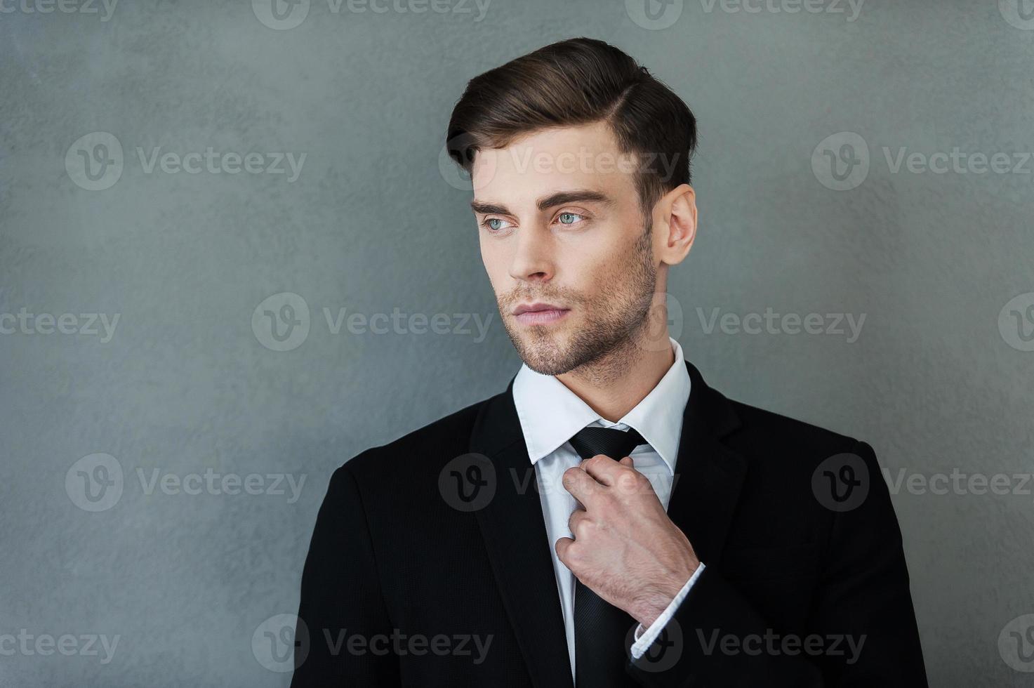 Confident businessman. Confident young businessman adjusting his necktie and looking away while standing against grey background photo