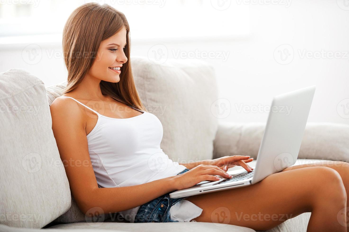 Lazy day with my laptop. Smiling young woman working on laptop while sitting on sofa photo