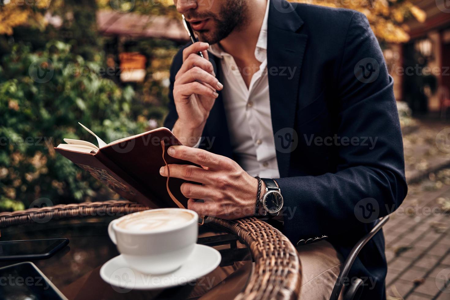 Busy day. Close up of young man in smart casual wear checking notes in personal organizer while sitting in restaurant outdoors photo