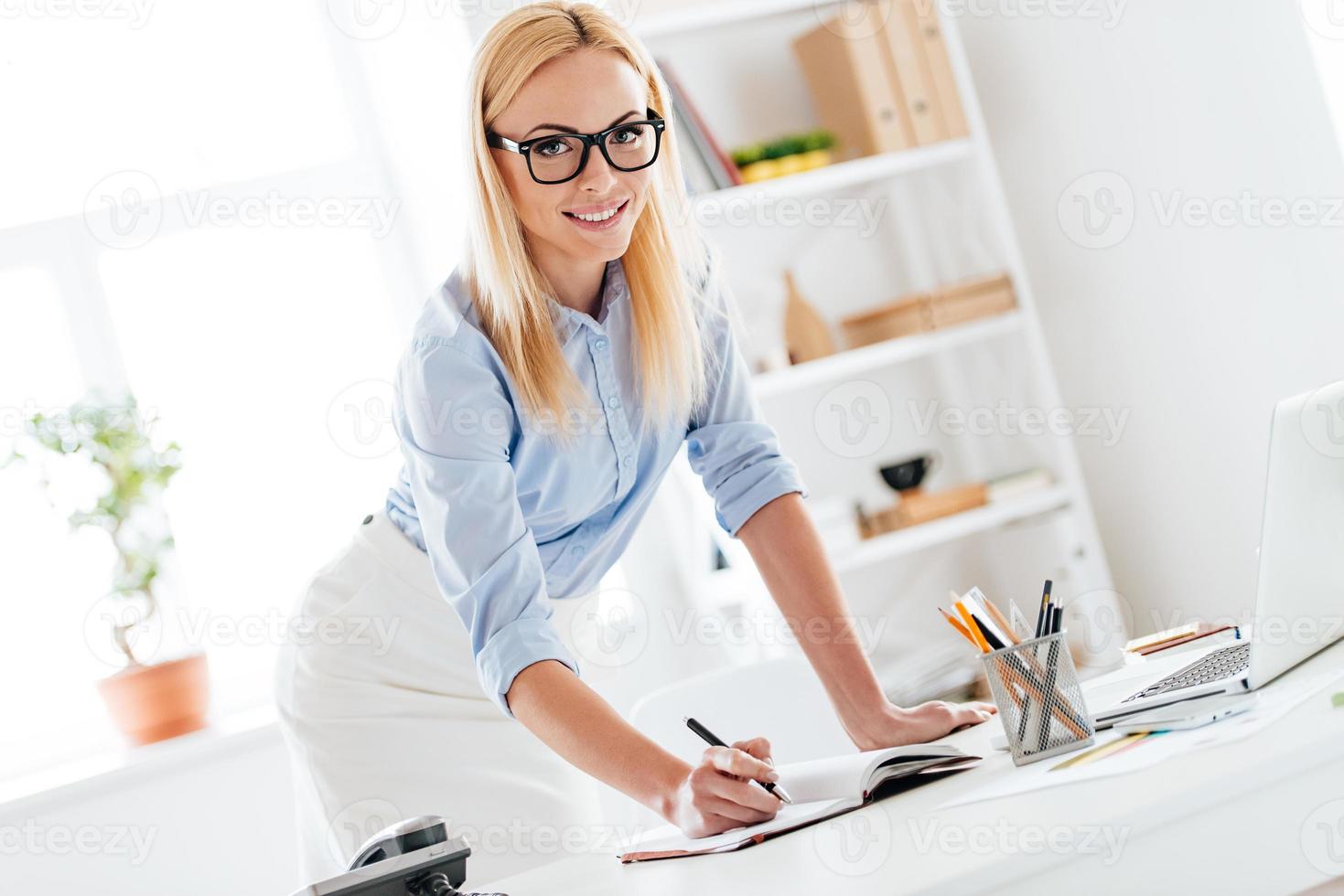 déjame escribirlo hermosa mujer tomando algunas notas y mirando a la cámara con una sonrisa mientras se inclina a la mesa en la oficina foto