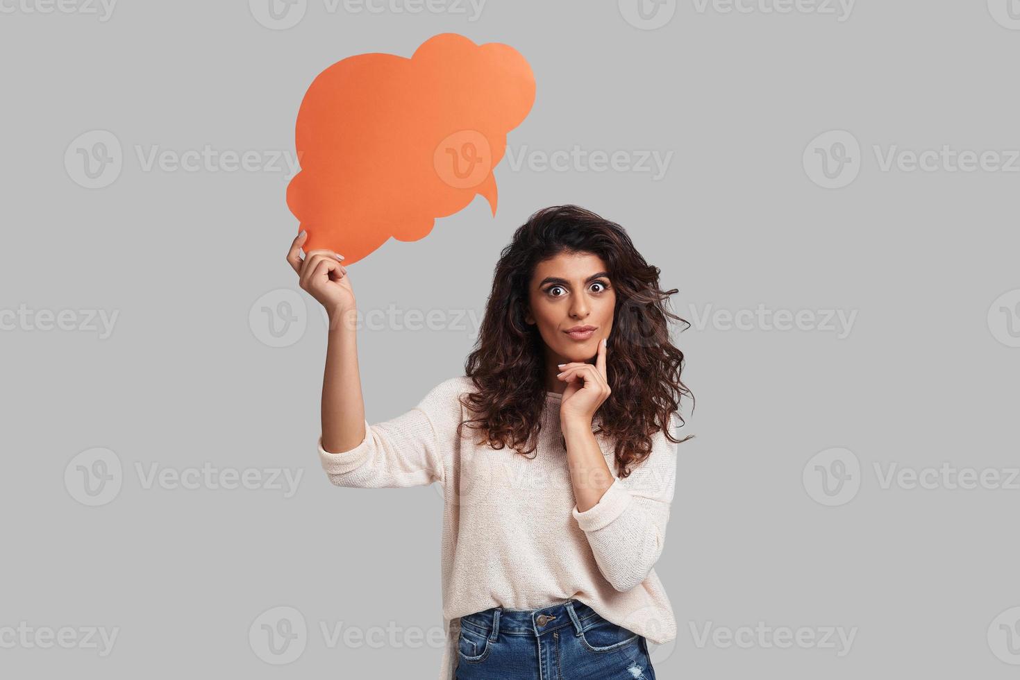 This is an idea Thoughtful young woman staring at camera and touching her face with index finger while standing against grey background photo