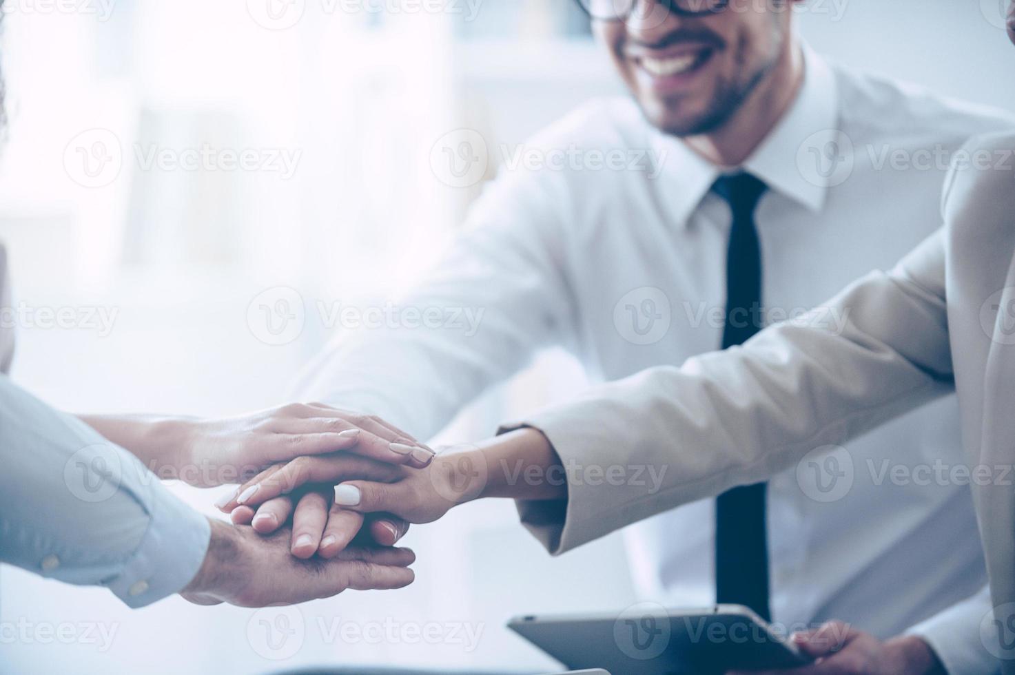 We are real team Close-up part of top view of group of four young people holding hands and showing their unity with smile while sitting at office photo