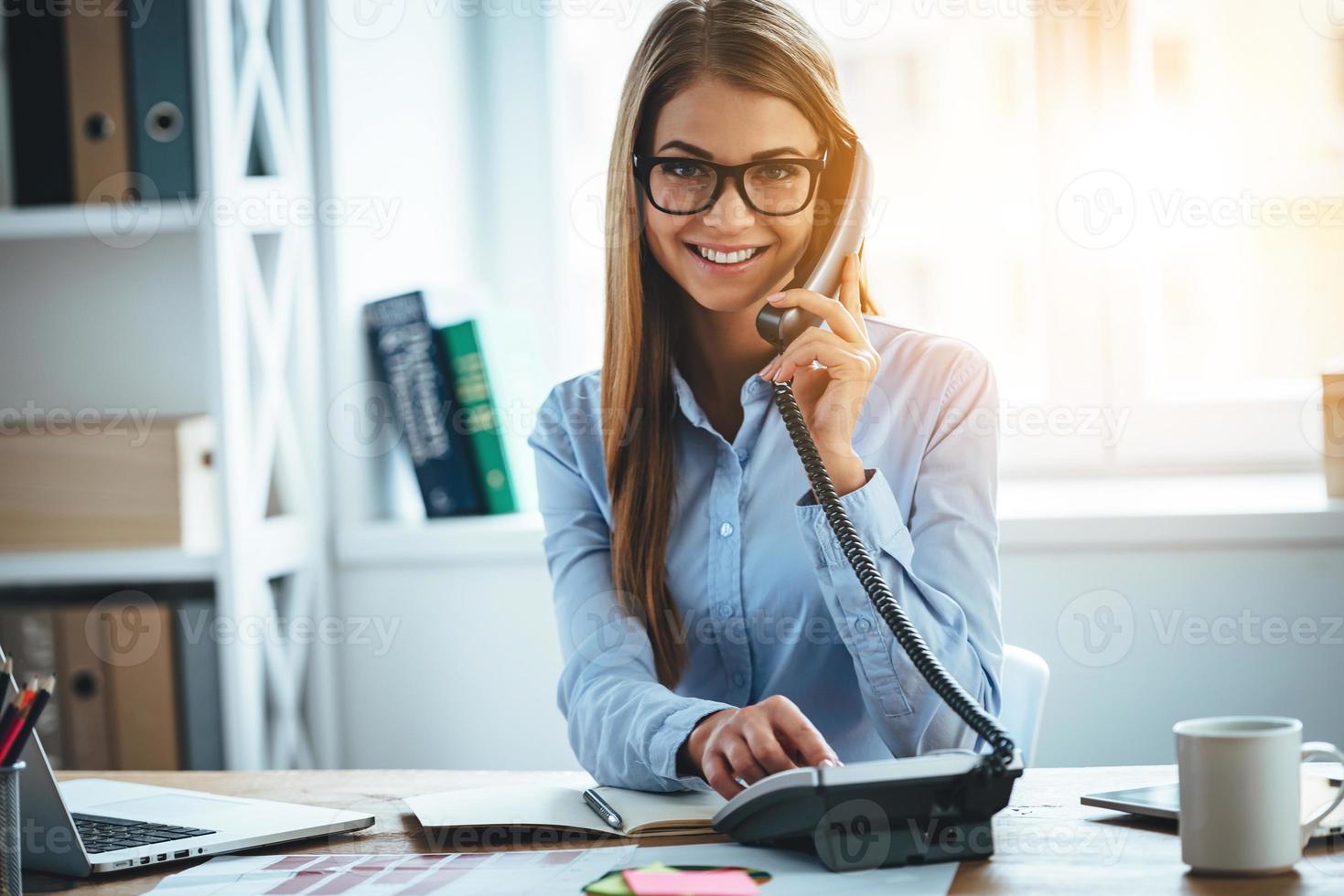 I will connect you in one second Cheerful young beautiful woman in glasses talking on the phone and looking at camera with smile while sitting at her working place photo