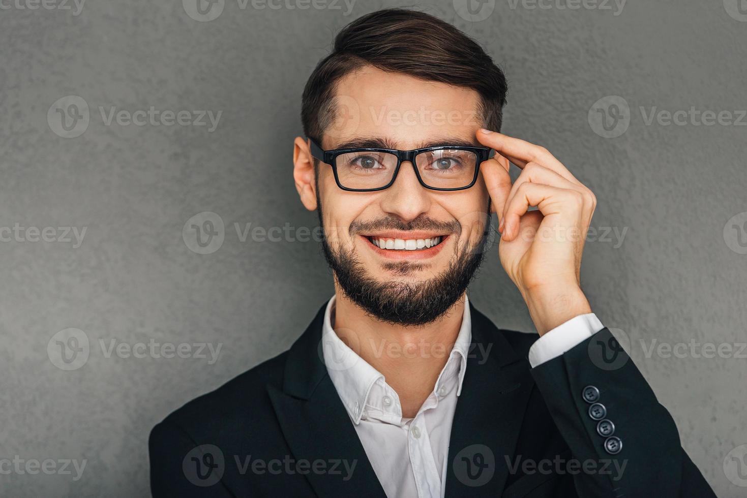 I clearly can see this Portrait of cheerful young man looking at camera with smile and adjusting his glasses while standing against grey background photo