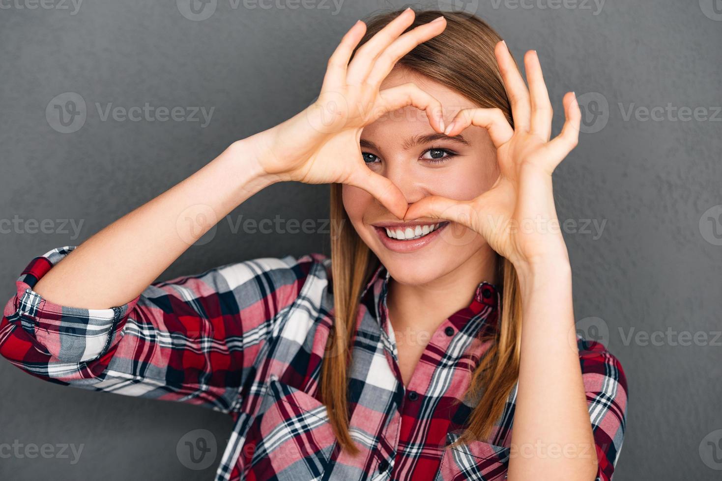 Love you Beautiful young woman making heart shape with her hands and looking at camera with smile while standing against grey background photo
