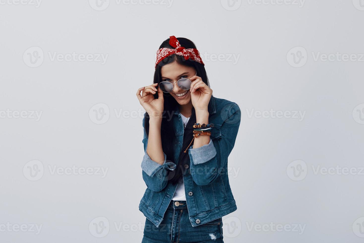 Attractive young woman in bandana adjusting eyewear and smiling while standing against grey background photo