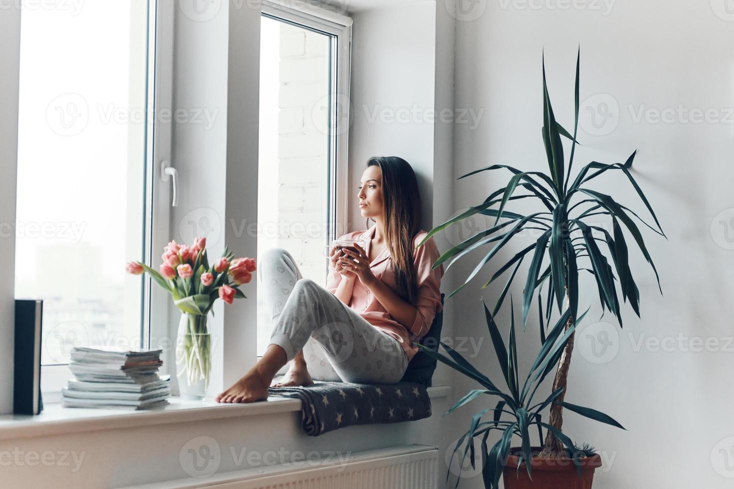 Thoughtful young woman in cozy pajamas looking through the window while resting at home photo