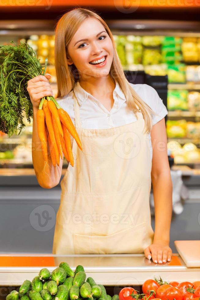 Vitamins for the customers. Cheerful young female seller smiling at camera and holding carrots while standing at a food store photo
