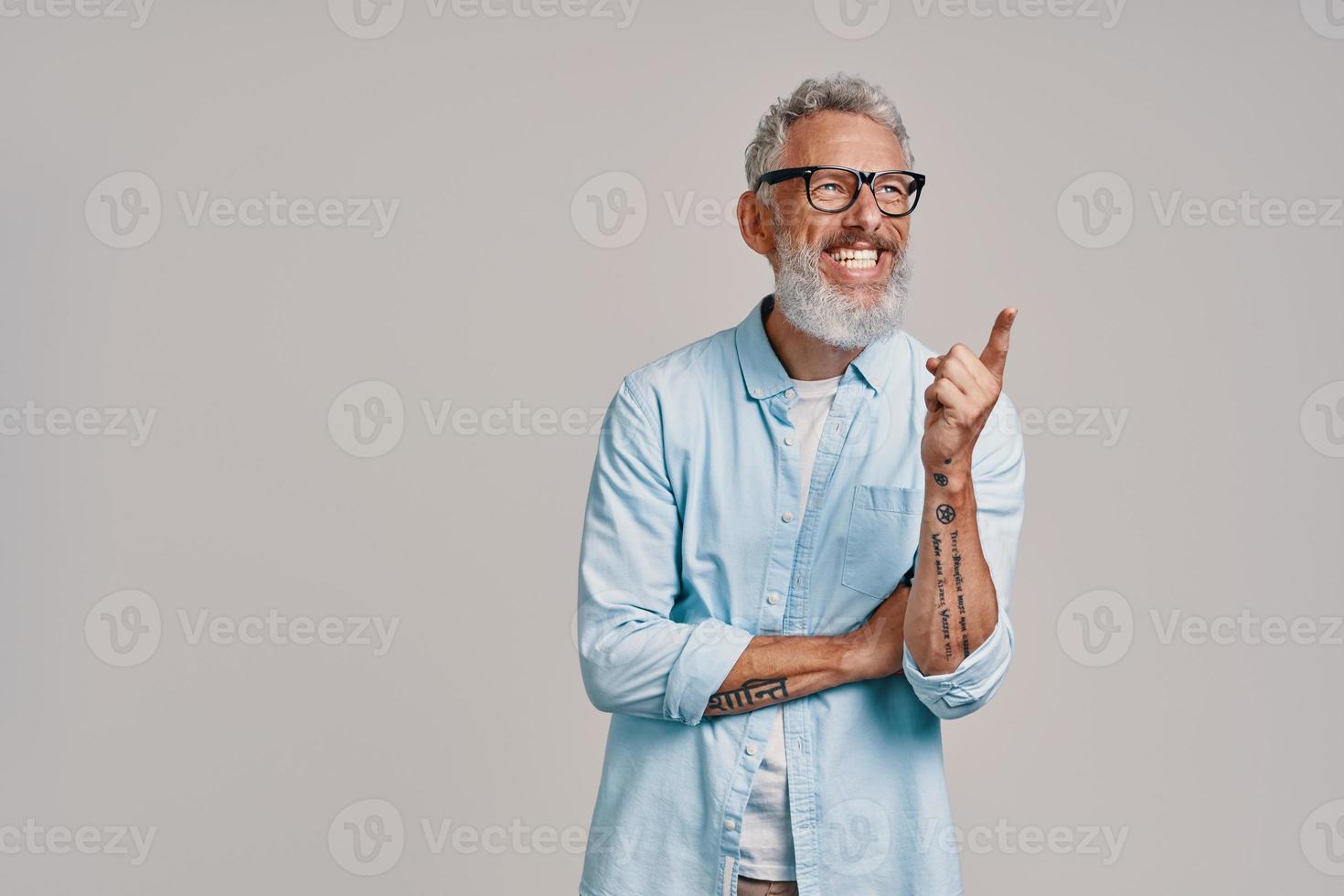 Handsome senior man pointing away and smiling while standing against gray background photo