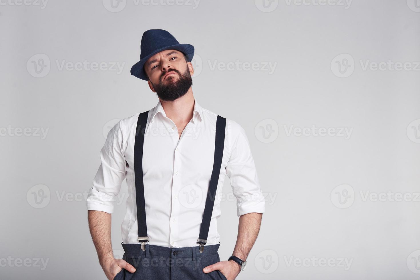 criminal elegante. un joven apuesto con tirantes manteniendo las manos en los bolsillos mirando a la cámara mientras se enfrenta a un fondo gris foto