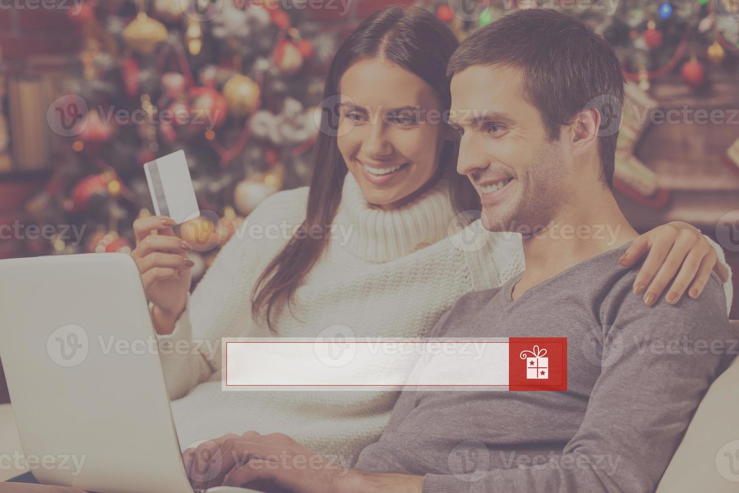 Find presents online. Beautiful young couple buying online while using computer together with Christmas Tree in the background photo