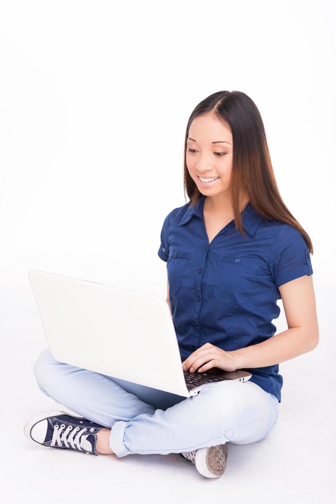 Woman surfing the net. Cheerful young Asian woman working on laptop and smiling while sitting against white background photo