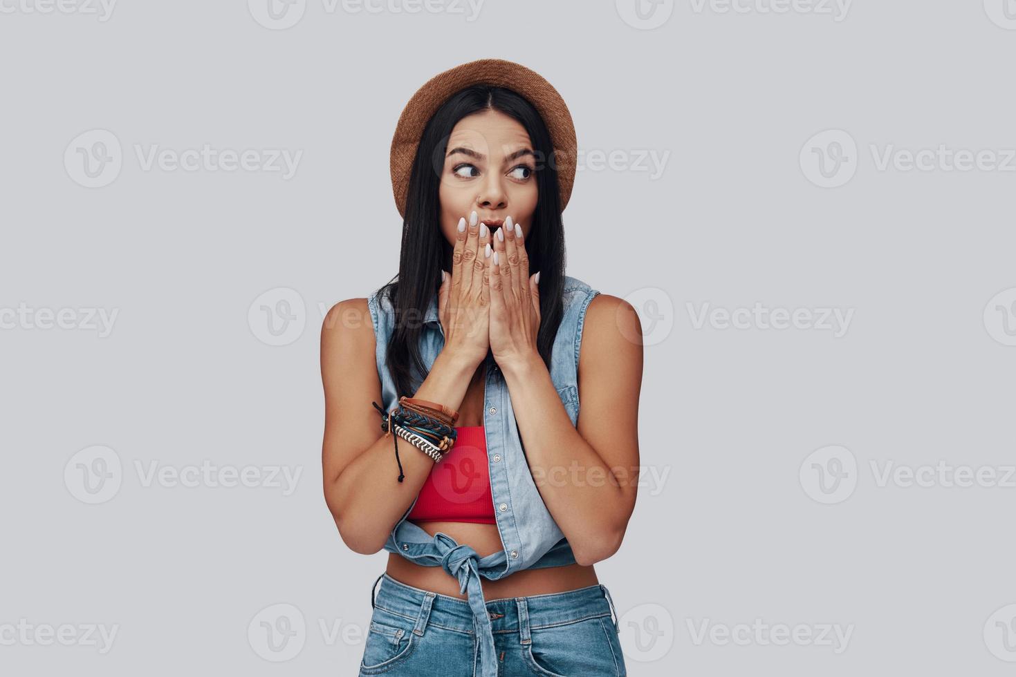 Surprised stylish young woman looking away and covering mouth with hands while standing against grey background photo