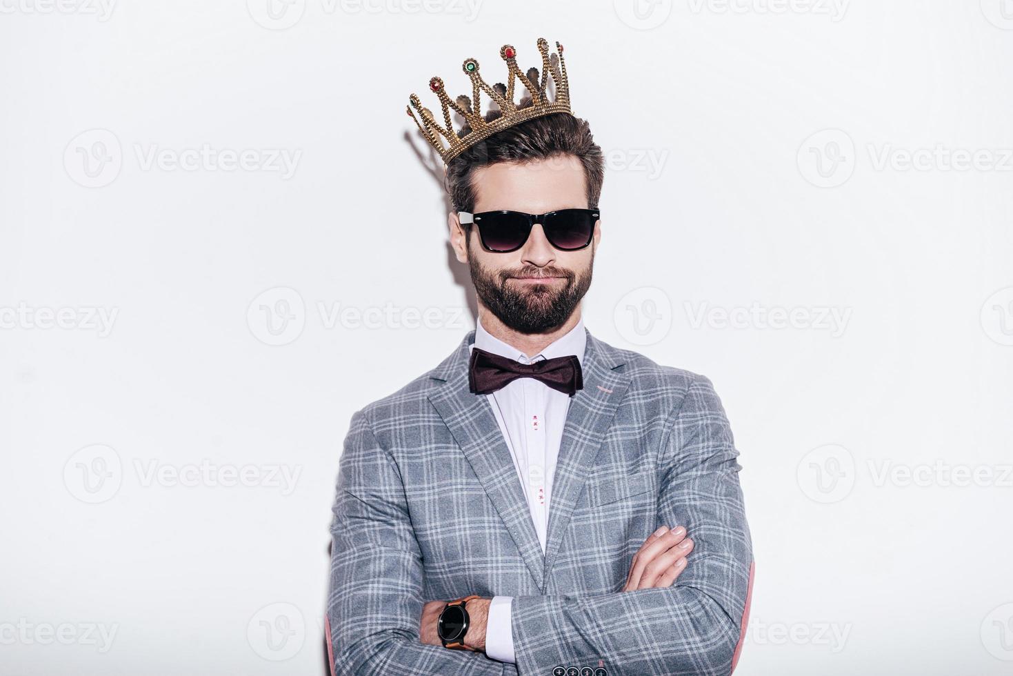 King of style. Sneering young handsome man wearing suit and crown keeping arms crossed and looking at camera while standing against white background photo