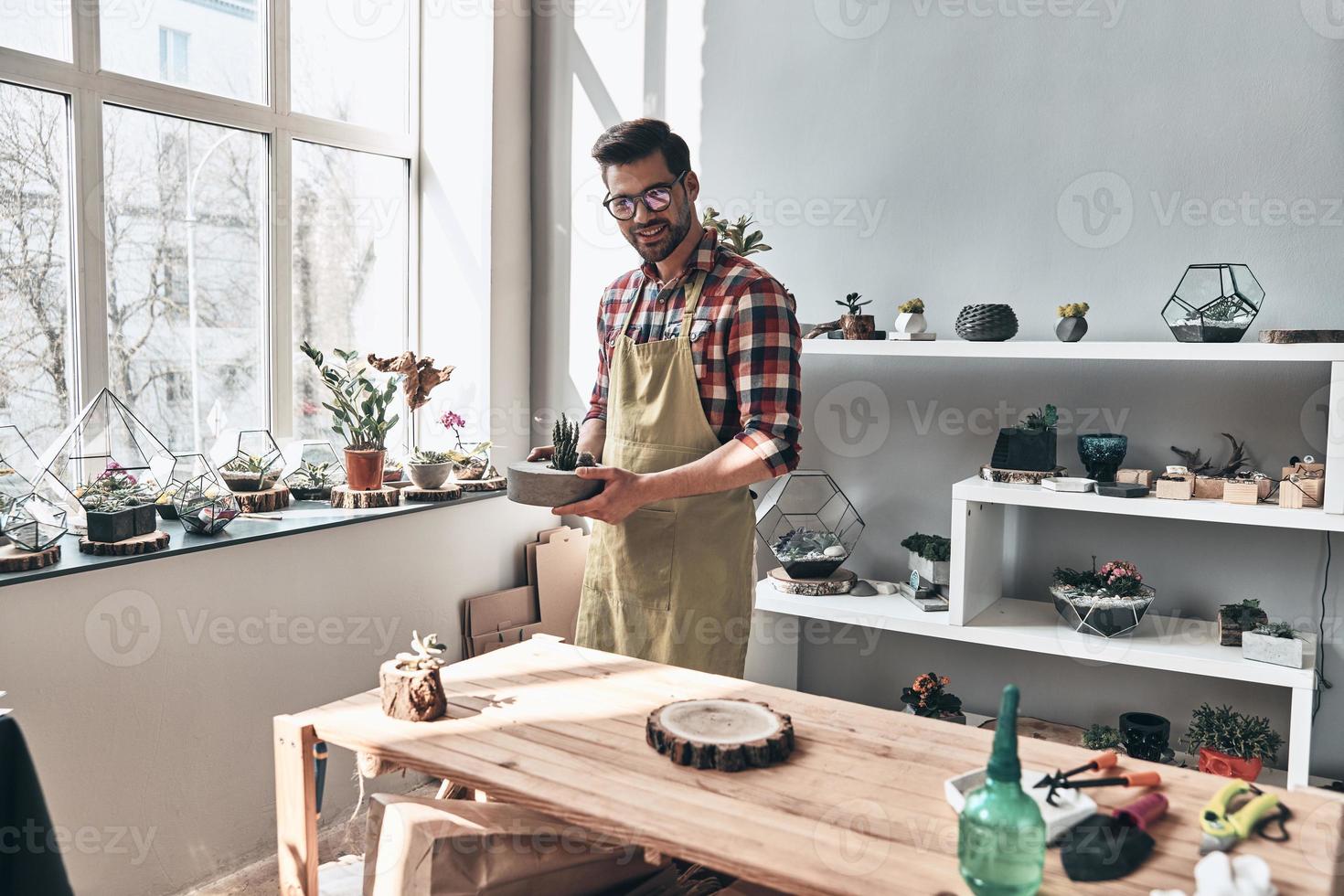 He turned his hobby into business. Handsome young man in apron looking at camera and smiling while standing in small garden center photo