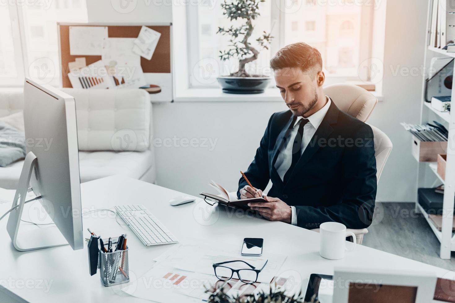 Making some notes. Handsome young man in full suit writing something in his notebook while sitting at the office desk photo