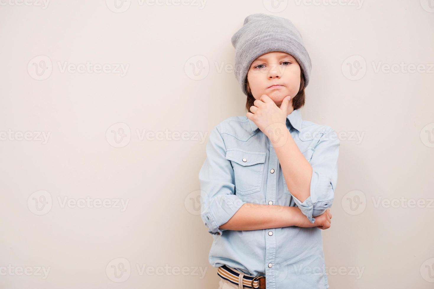 Deep in his thoughts. Handsome little boy in hat holding hand on chin and looking at camera while standing against grey background photo