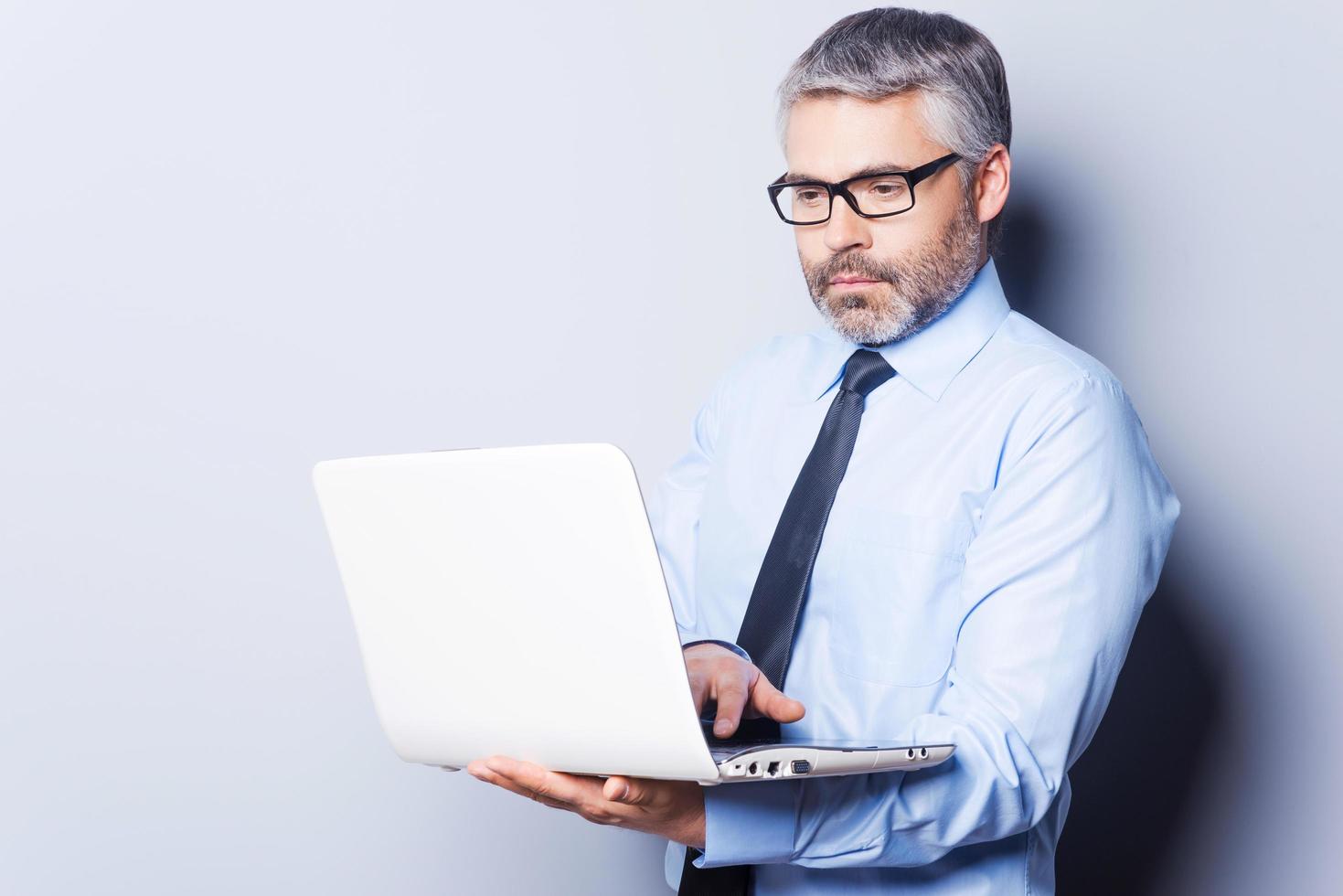 Supporting your business. Confident mature man in formalwear working on laptop while standing isolated on white background photo