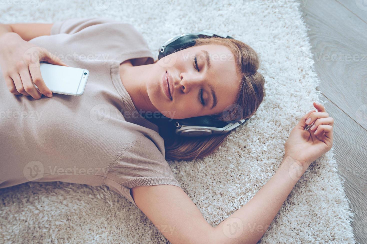 Feeling calm. Top view of beautiful young woman listening to music and keeping eyes closed while lying on carpet at home photo