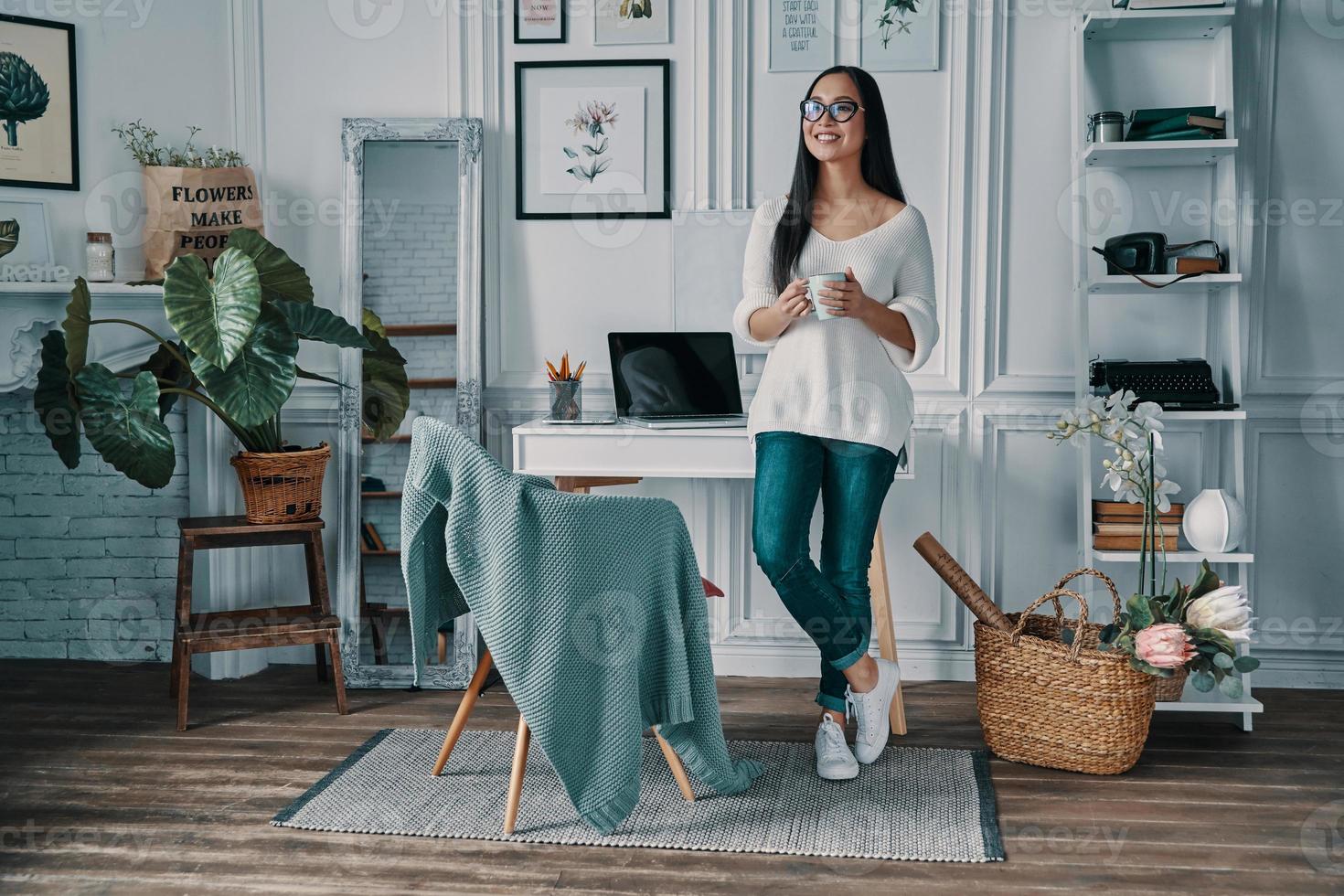 Enjoying fresh coffee. Full length of beautiful young woman looking away and smiling while standing in home office photo