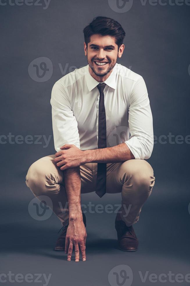 ¿Puedes resistirte a su sonrisa completa de un joven apuesto y seguro de sí mismo mirando a la cámara con una sonrisa mientras te sientas agachado contra un fondo gris? foto