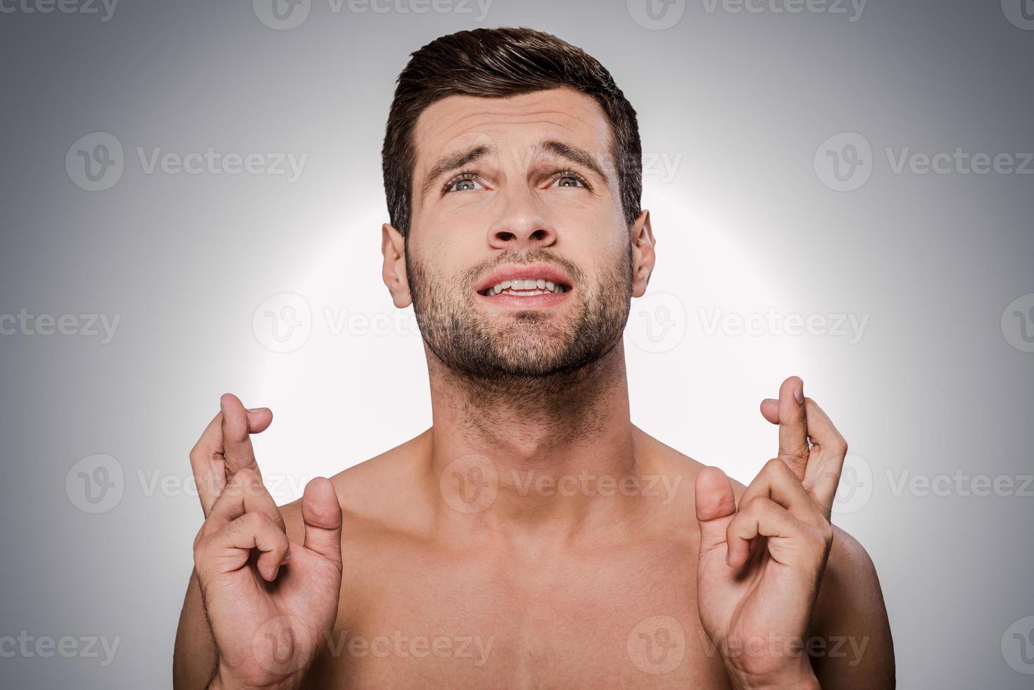 Begging for... Portrait of young shirtless man keeping fingers crossed and looking up while standing against grey background photo