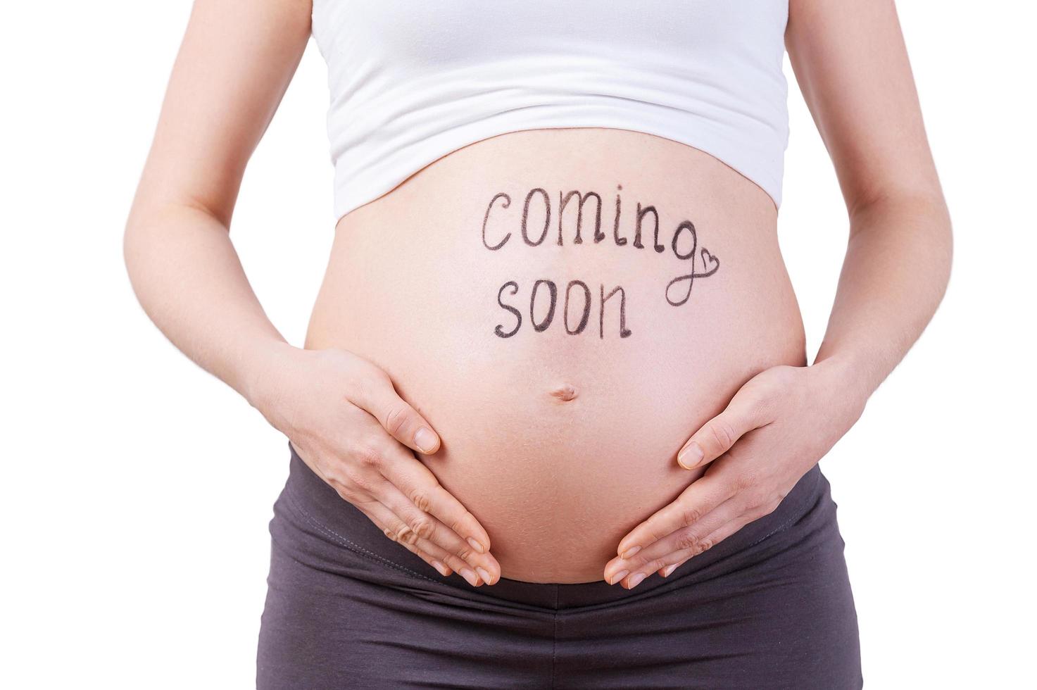 Coming soon. Cropped image of pregnant woman with coming soon sign on her belly standing isolated on white photo