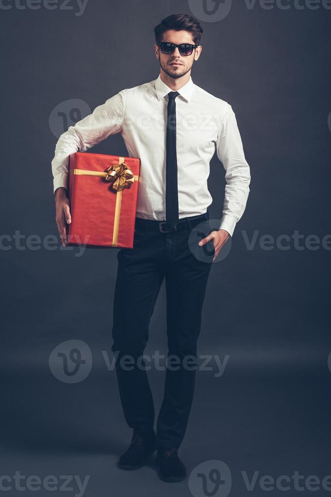 Surprise for you. Full length of handsome well-dressed young man in sunglasses holding gift box and looking at camera while standing against grey background photo