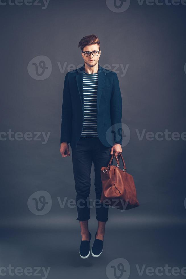 Style in mid air. Full length of confident young handsome man in glasses carrying bag and looking at camera while jumping against grey background photo