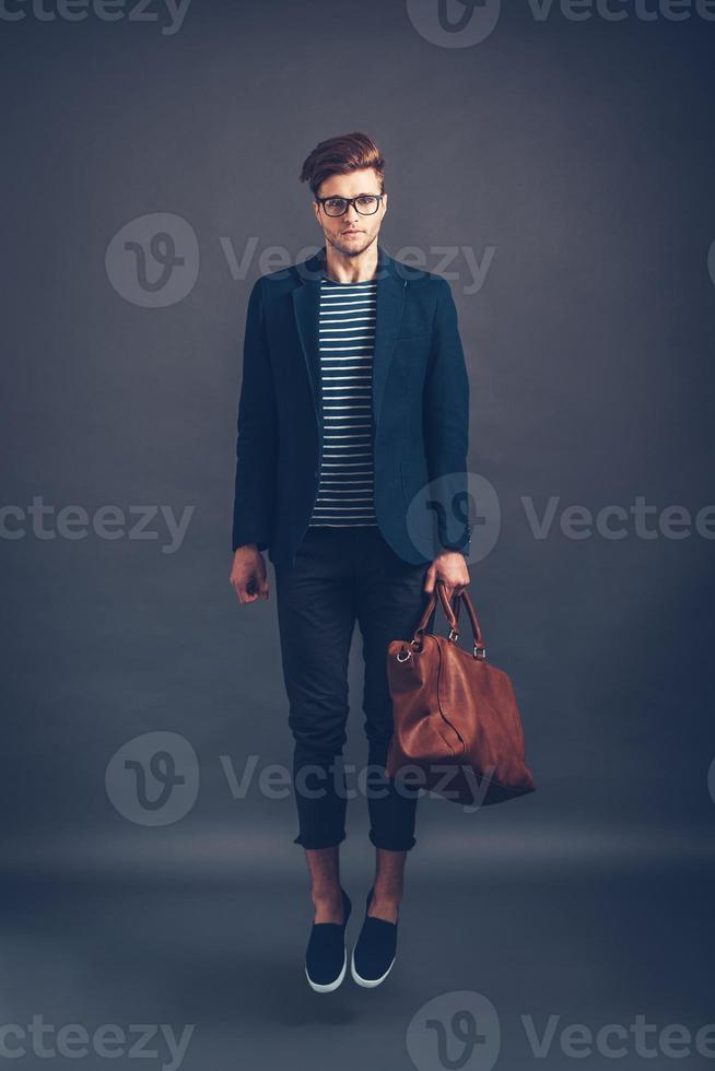 estilo en el aire. toda la longitud de un joven apuesto y seguro de sí mismo con gafas que lleva una bolsa y mira la cámara mientras salta contra un fondo gris foto