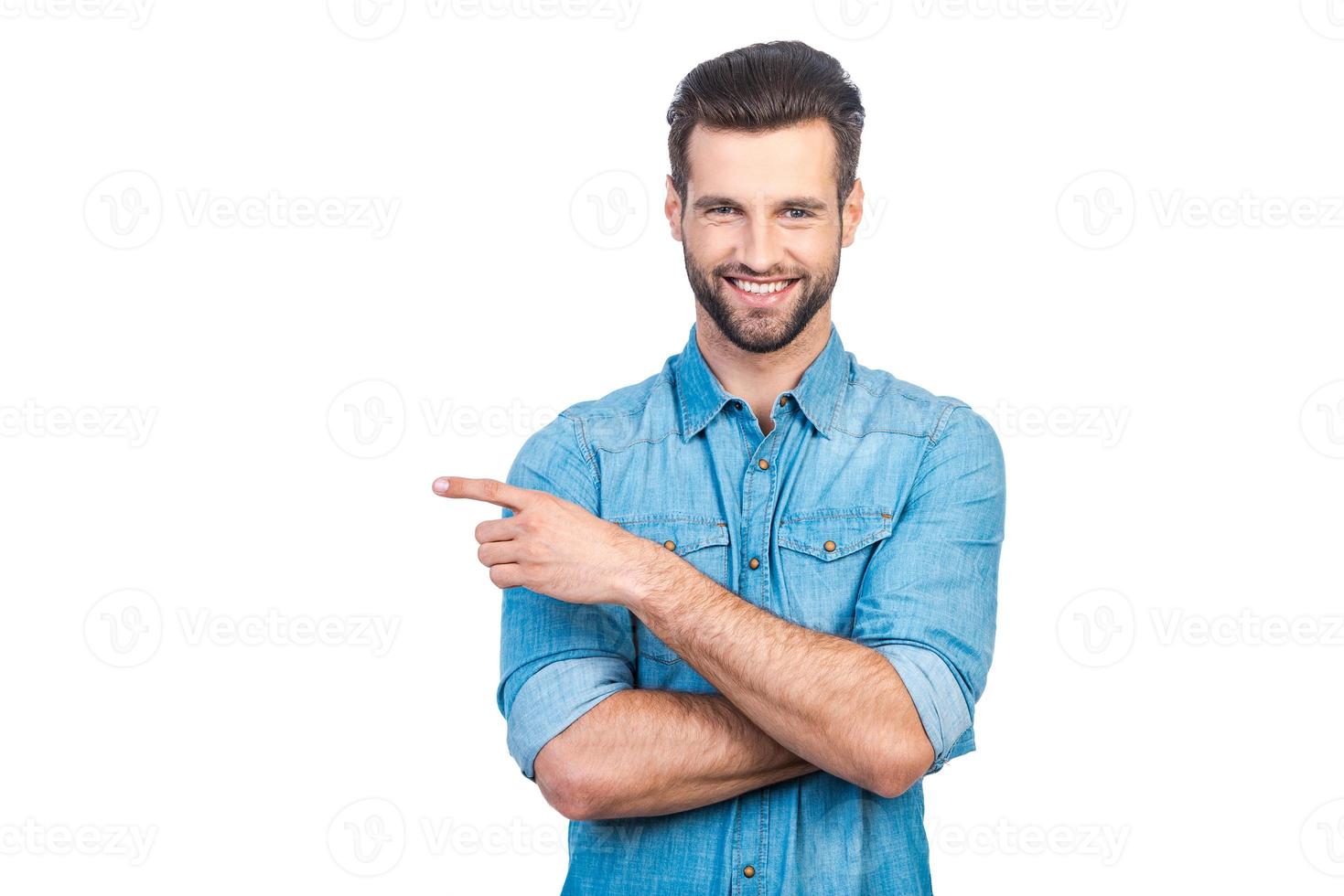 mire hacia allí un joven apuesto feliz con camisa de jeans apuntando hacia otro lado y sonriendo mientras está de pie contra el fondo blanco foto