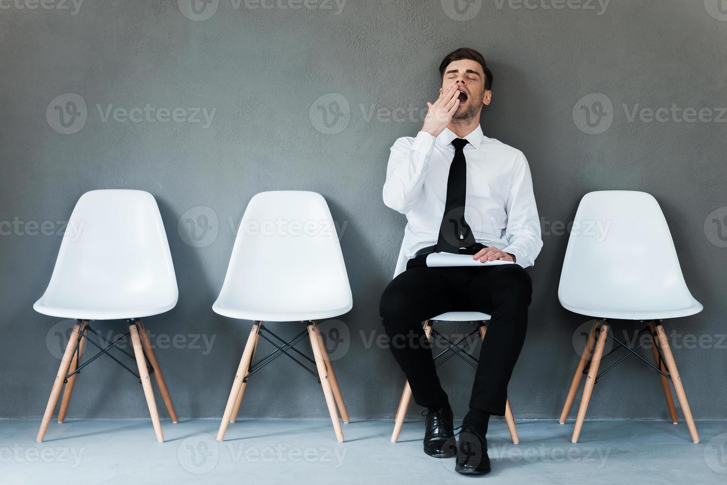 Tired of waiting. Tired young businessman holding paper and yawning while sitting on chair against grey background photo