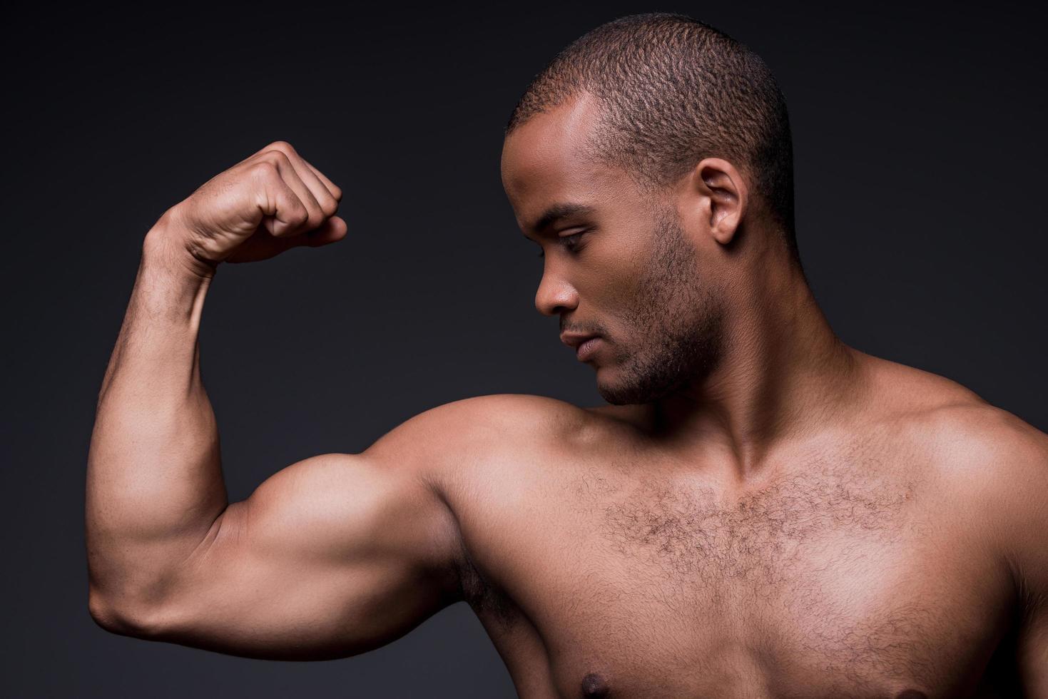 orgulloso de sus bíceps perfectos. joven africano sin camisa mirando sus bíceps mientras está de pie contra el fondo negro foto