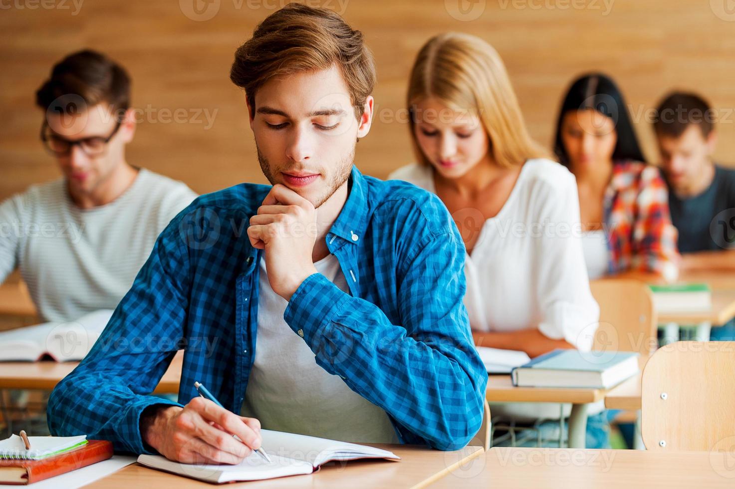 Focused on exam. Group of concentrated young students writing something in their note pads while sitting at their desks in the classroom photo