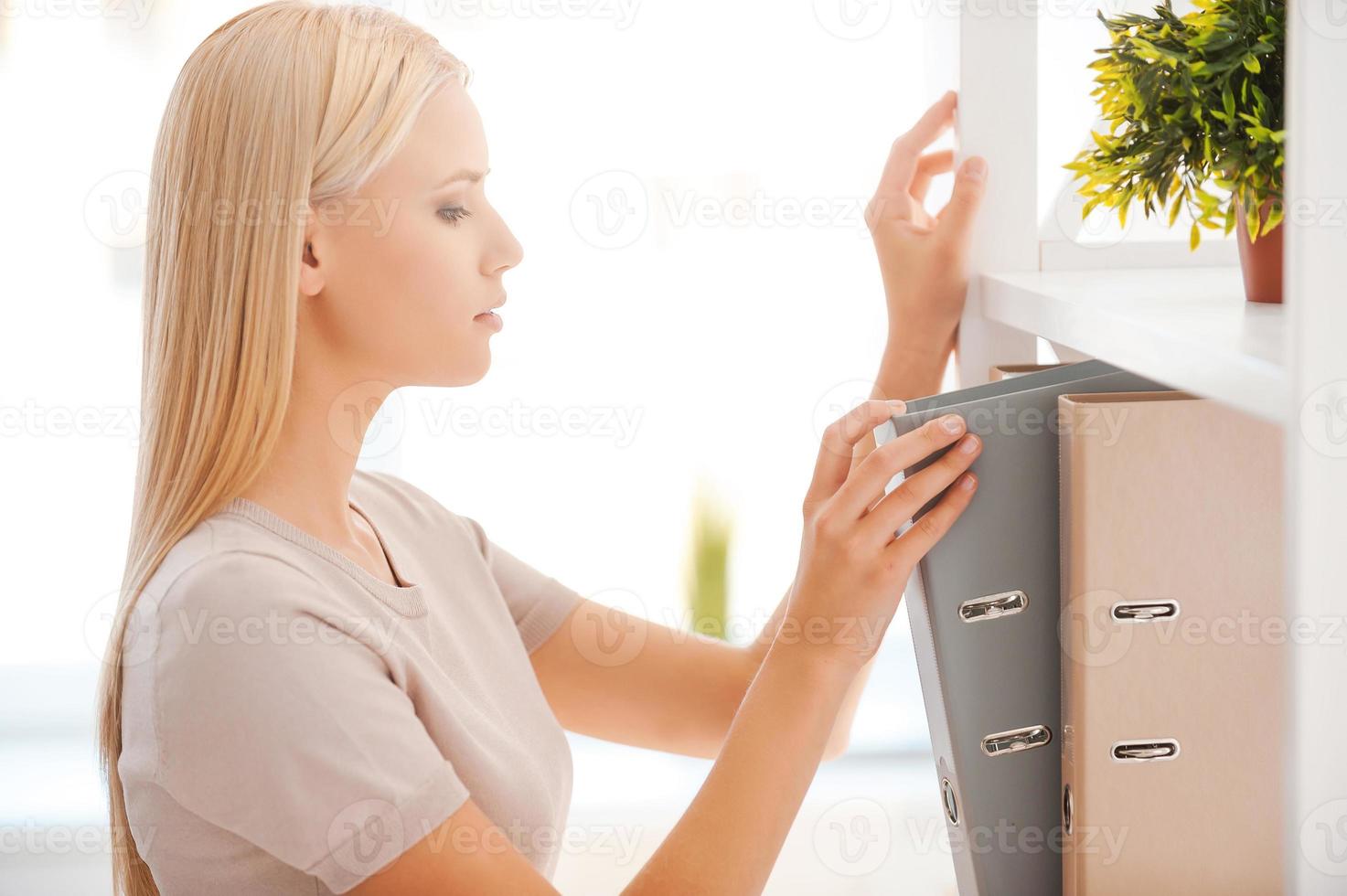 Looking for that document. Side view of beautiful young woman looking for the document she need while standing near the self in office photo