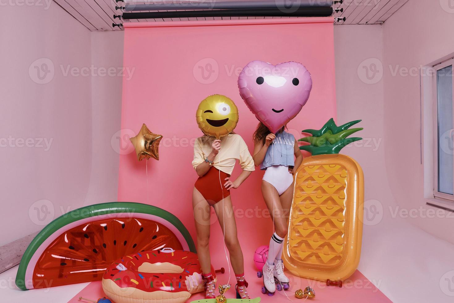 divirtiéndose. Longitud total de dos mujeres jóvenes en traje de baño cubriendo las caras con globos mientras están de pie contra el fondo rosa foto