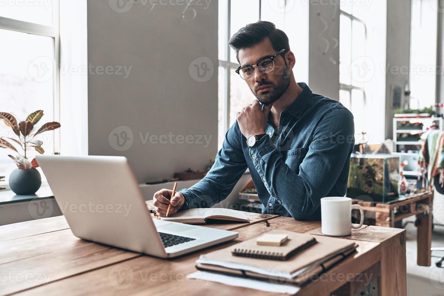 Achieving best results. Thoughtful young man keeping hand on chin and looking at camera while working in the creative working space photo